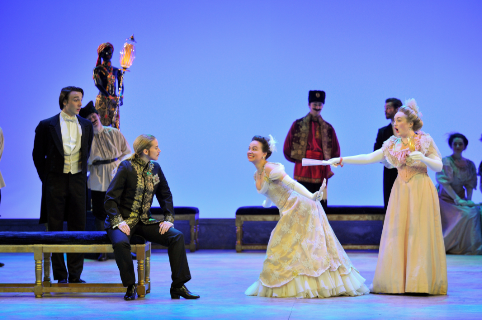 A group of students, dressed up in historical clothing, performing an opera, on stage, with other students dressed in historical clothing in the background, with a light blue background behind them.